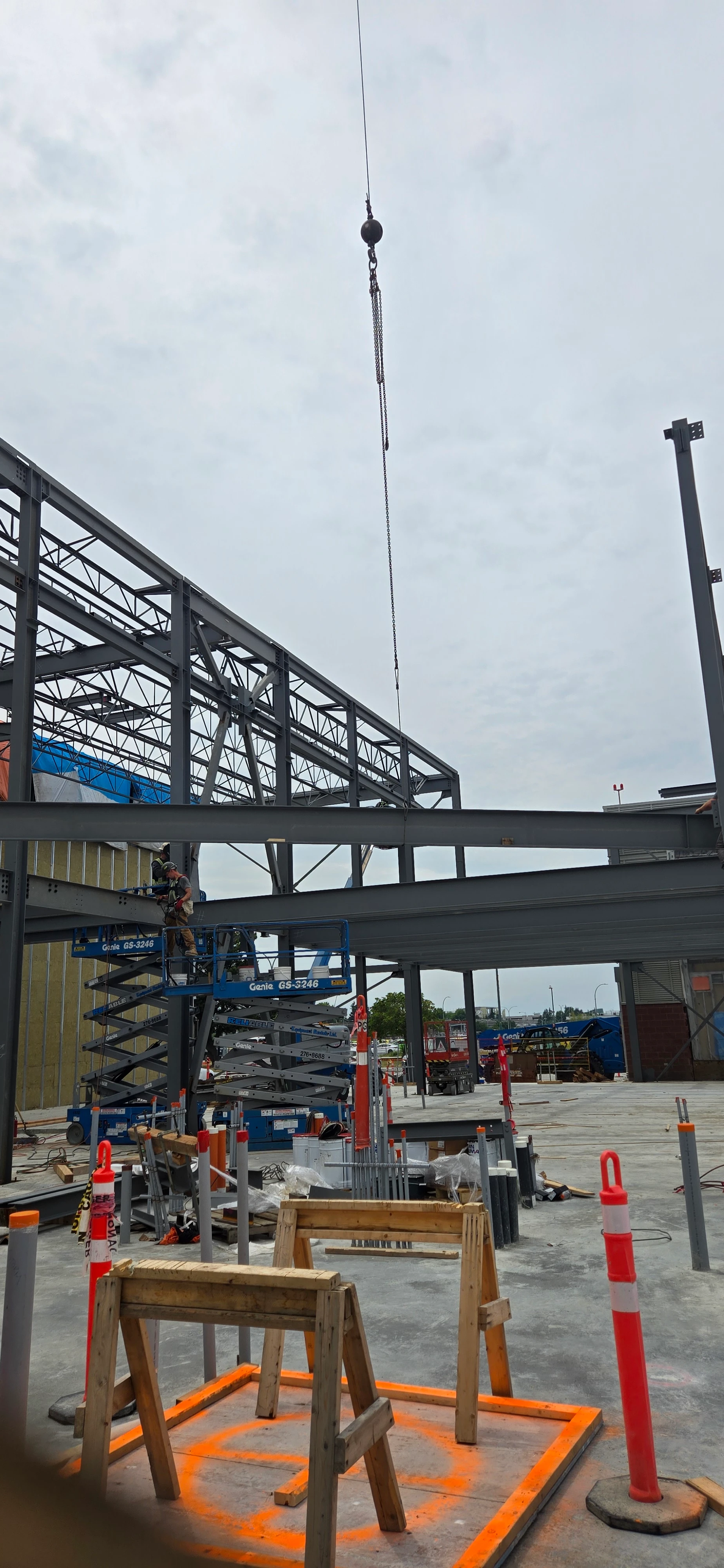 A view of a construction site focusing on the steel framework of a building. A crane hook is visible at the top, and various construction materials and safety cones are in the foreground.