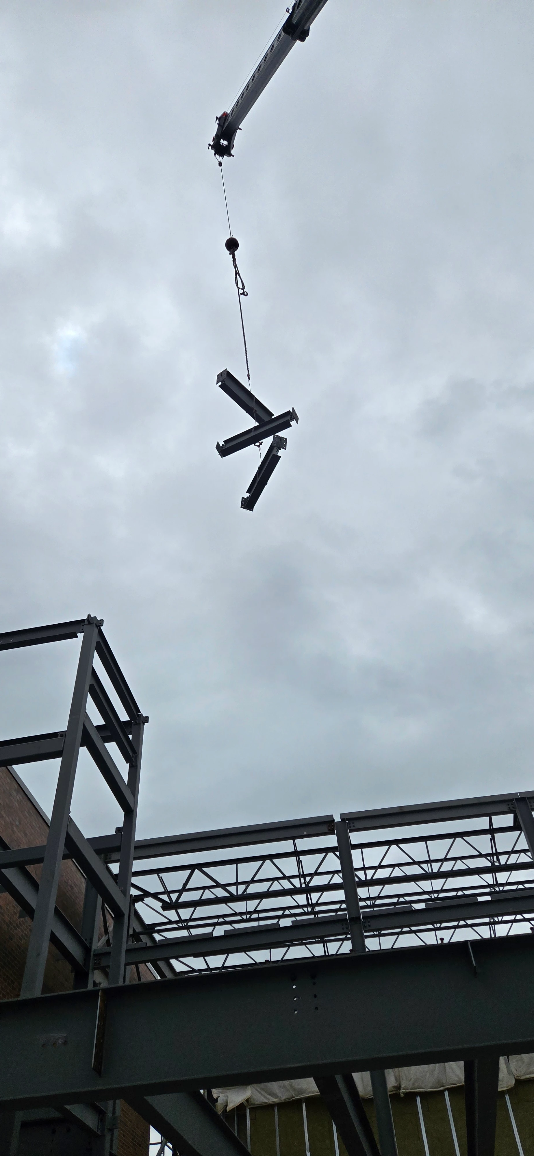 A crane hook lowering several short metal beams onto a construction site. The steel framework of a building is visible in the lower part of the image under a cloudy sky.