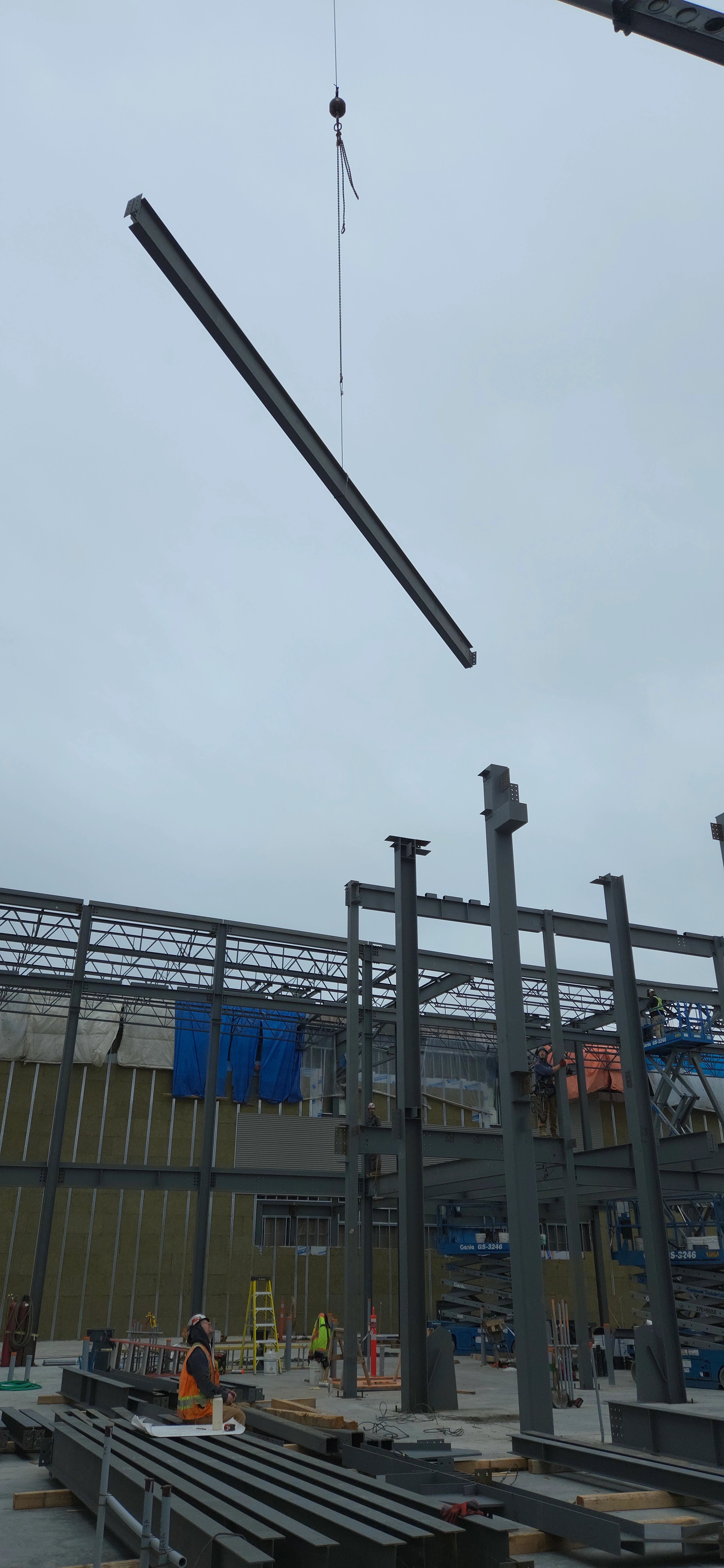 A large metal beam is being hoisted by a crane over a construction site. Workers are on the ground and on elevated platforms, and the steel structure of a building is taking shape.