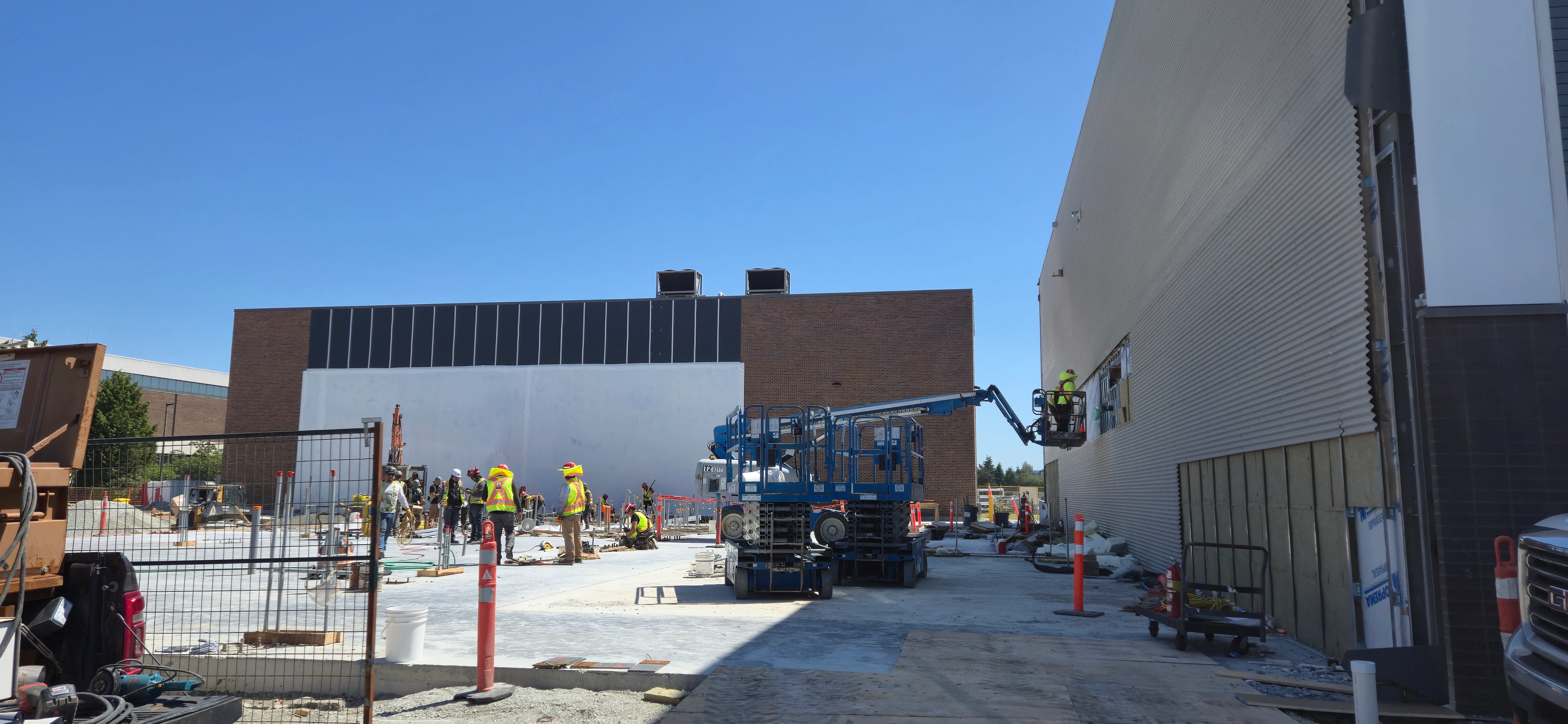 A wide shot of a construction site on a sunny day. Several workers in hard hats are on the ground, while one worker in a boom lift works on the side of a building with corrugated metal siding.