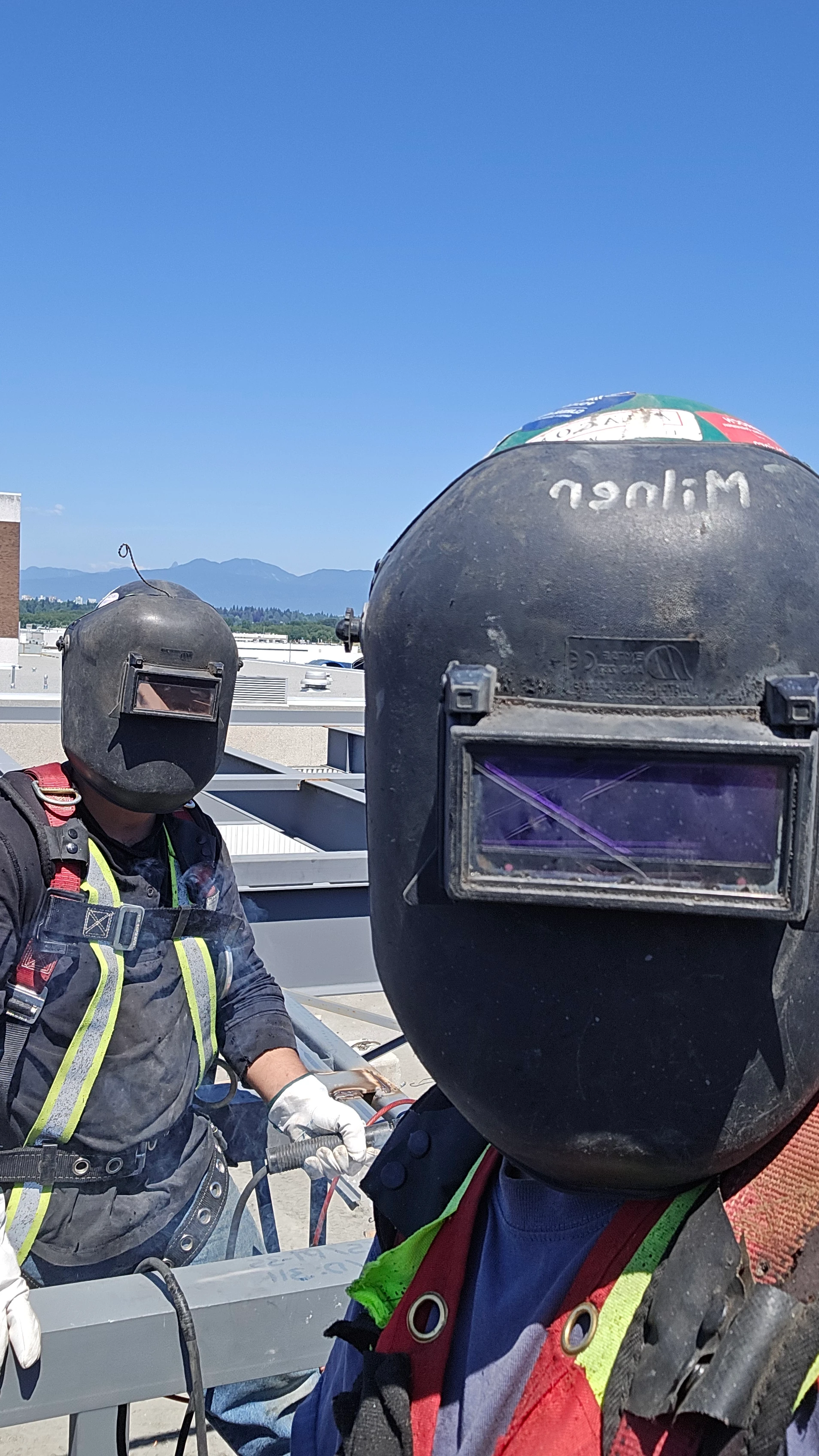 Two construction workers in welding helmets and safety harnesses, with one worker in the foreground and the other slightly behind and to the left. Mountains are visible in the distant background under a clear blue sky.