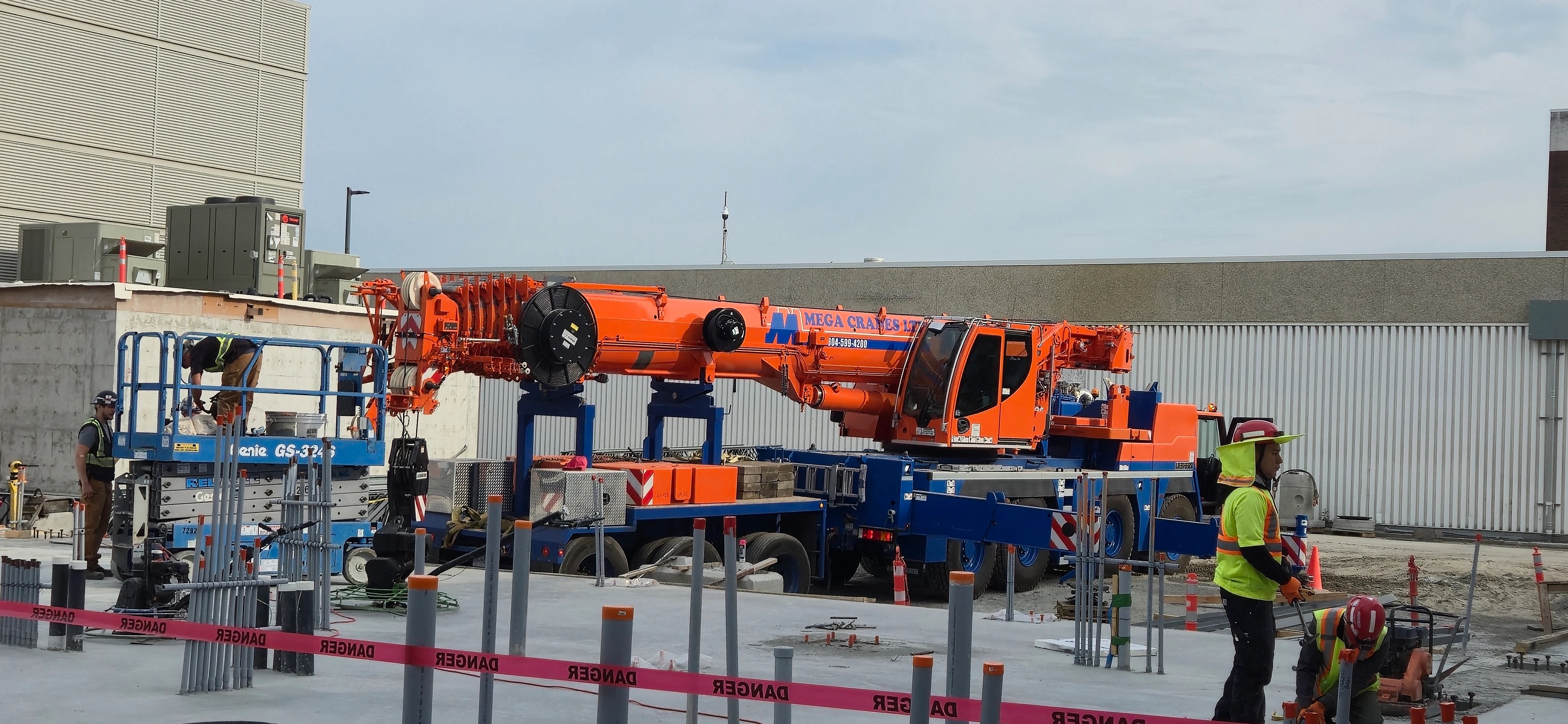 A large, orange and blue mobile crane is parked on a construction site. Workers are nearby, some on a scissor lift and others on the ground. Red "DANGER" tape is strung across the foreground.