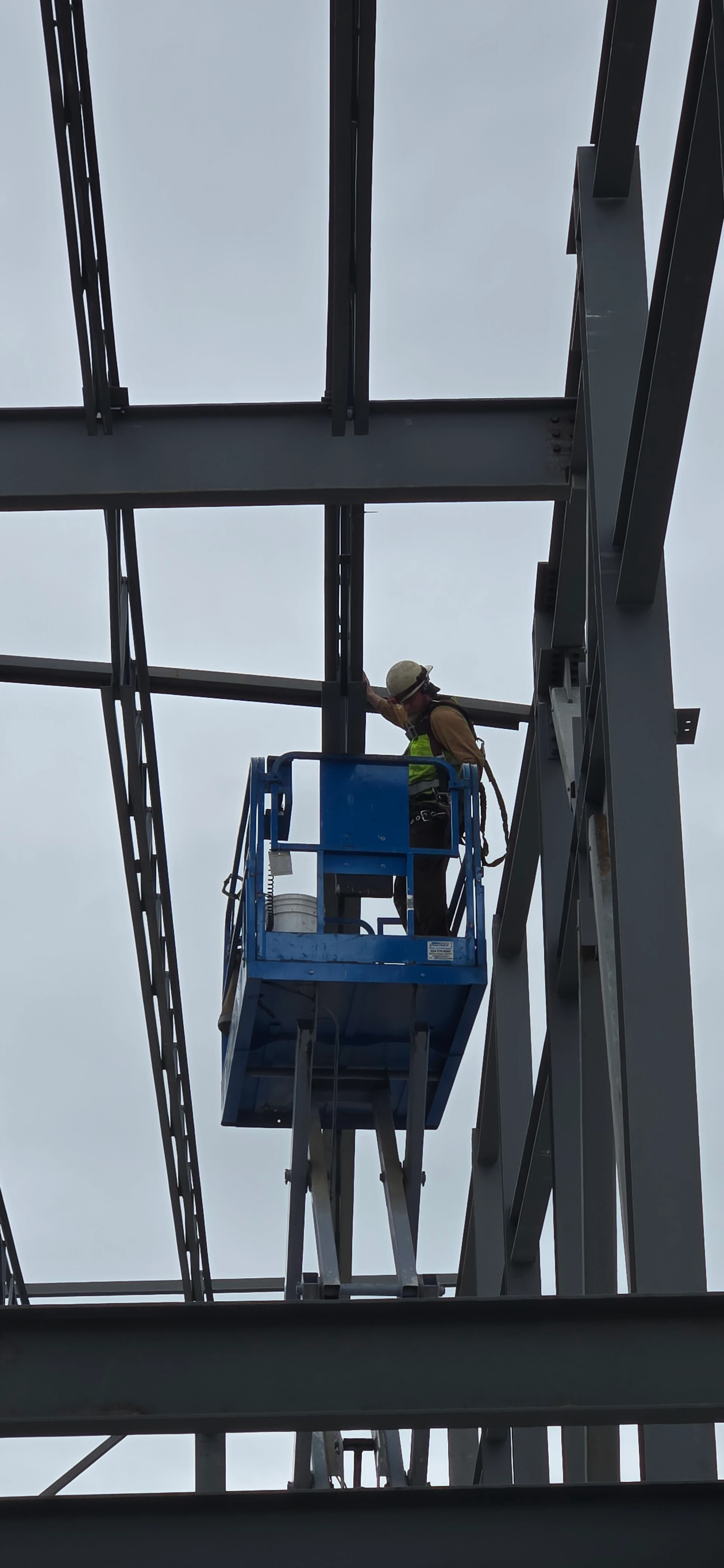 A low-angle shot of a construction worker in a hard hat and safety vest standing in a blue scissor lift. The worker is leaning towards a steel column within the building's large steel frame against an overcast sky.