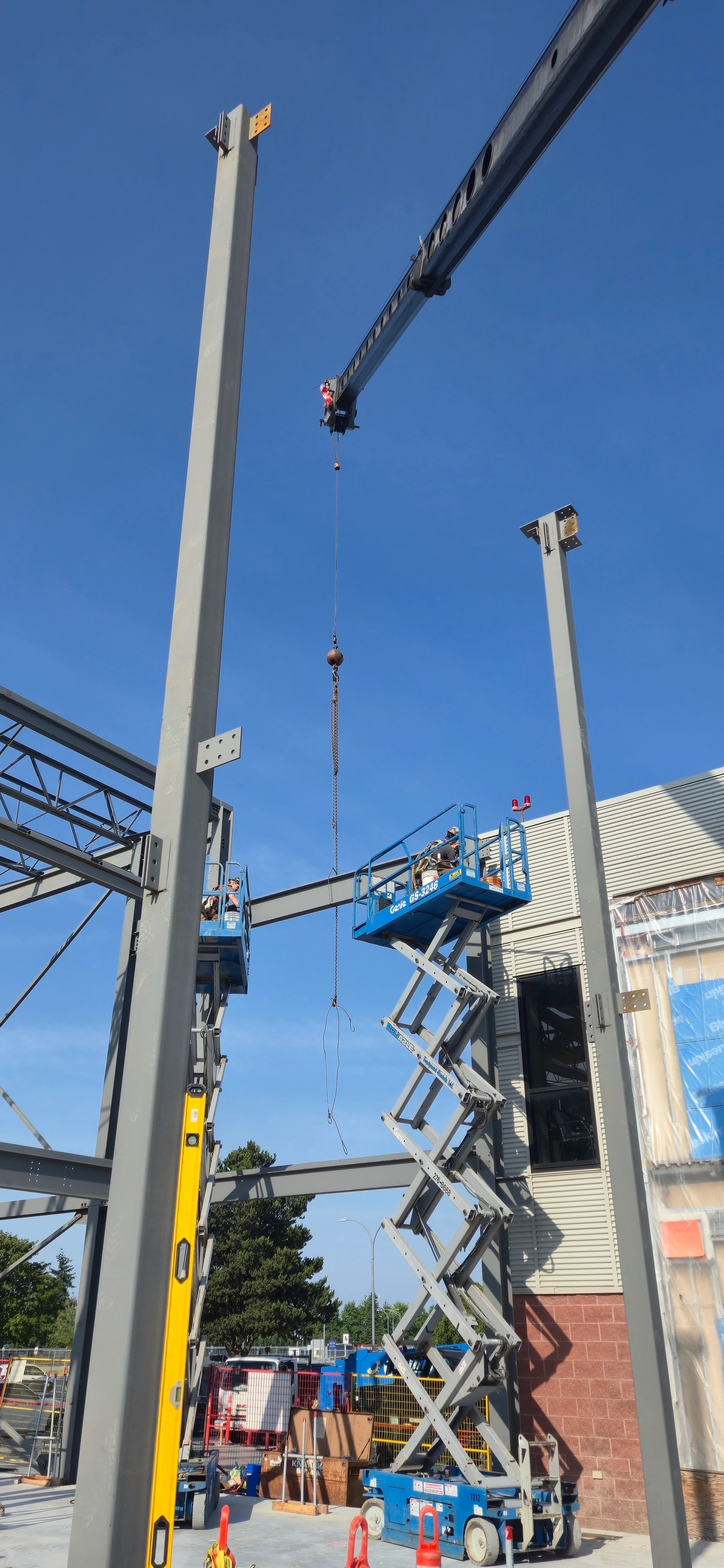 A clear blue sky above a construction site where a crane is lowering a beam. Two workers are on scissor lifts, positioned near the tall steel columns of the building framework.