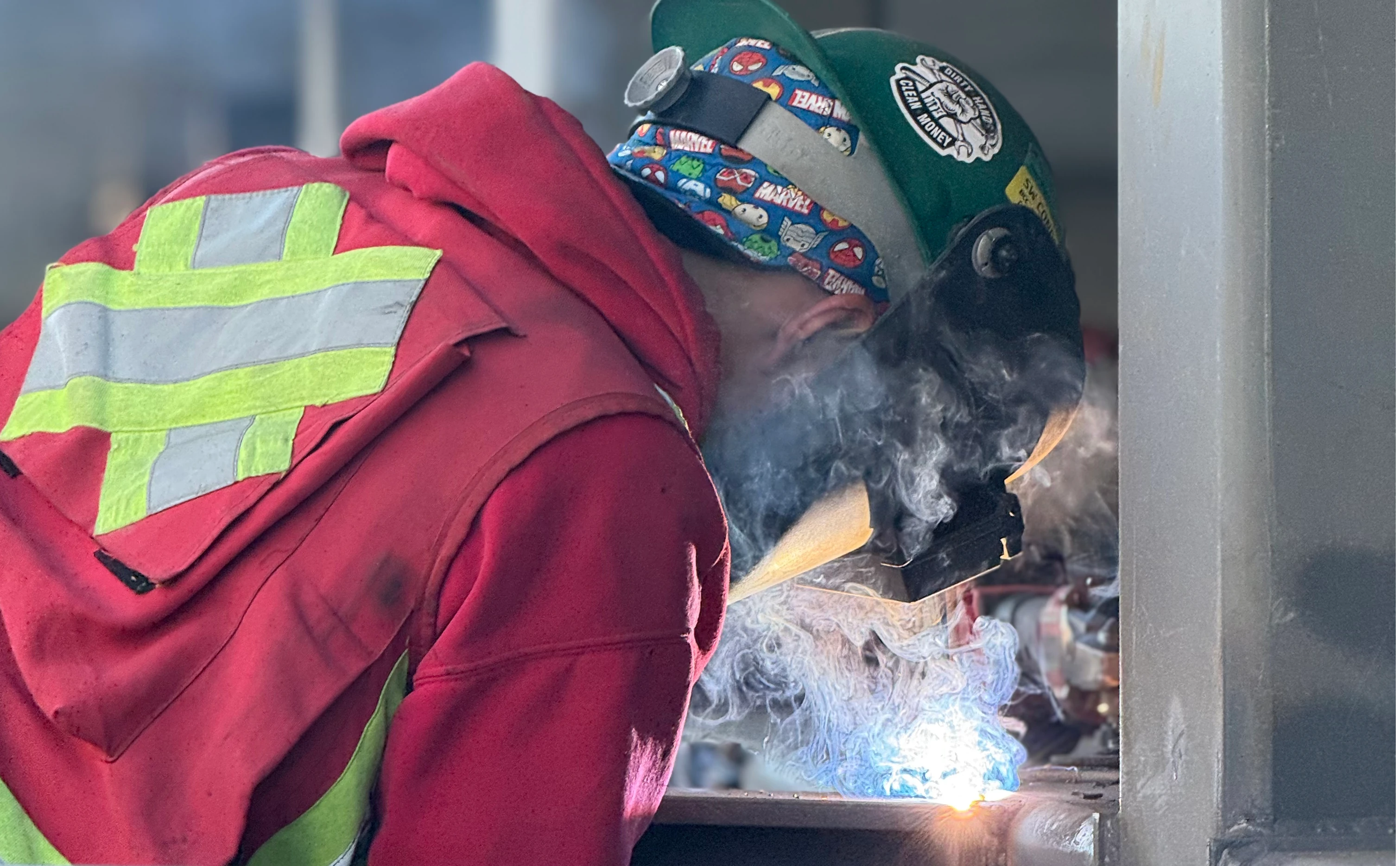 A welder wearing a red jacket, green hard hat, and protective hood is actively welding a metal piece, generating bright sparks and smoke. The welder's face is obscured by the hood.