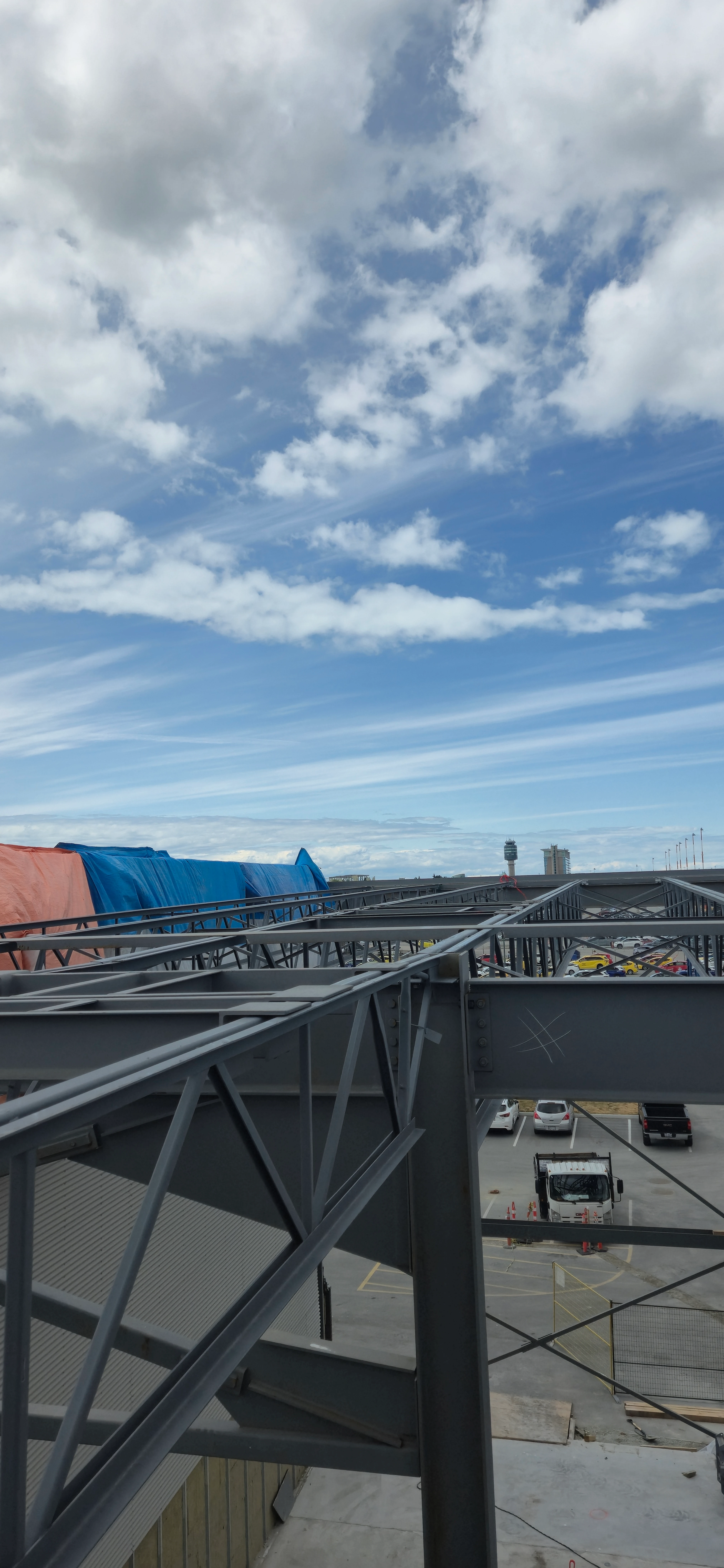 A high-angle view from a building's steel frame, looking out over a steel-trussed walkway. Below is a parking lot, and in the distance, an airport control tower is visible under a blue sky with white clouds.