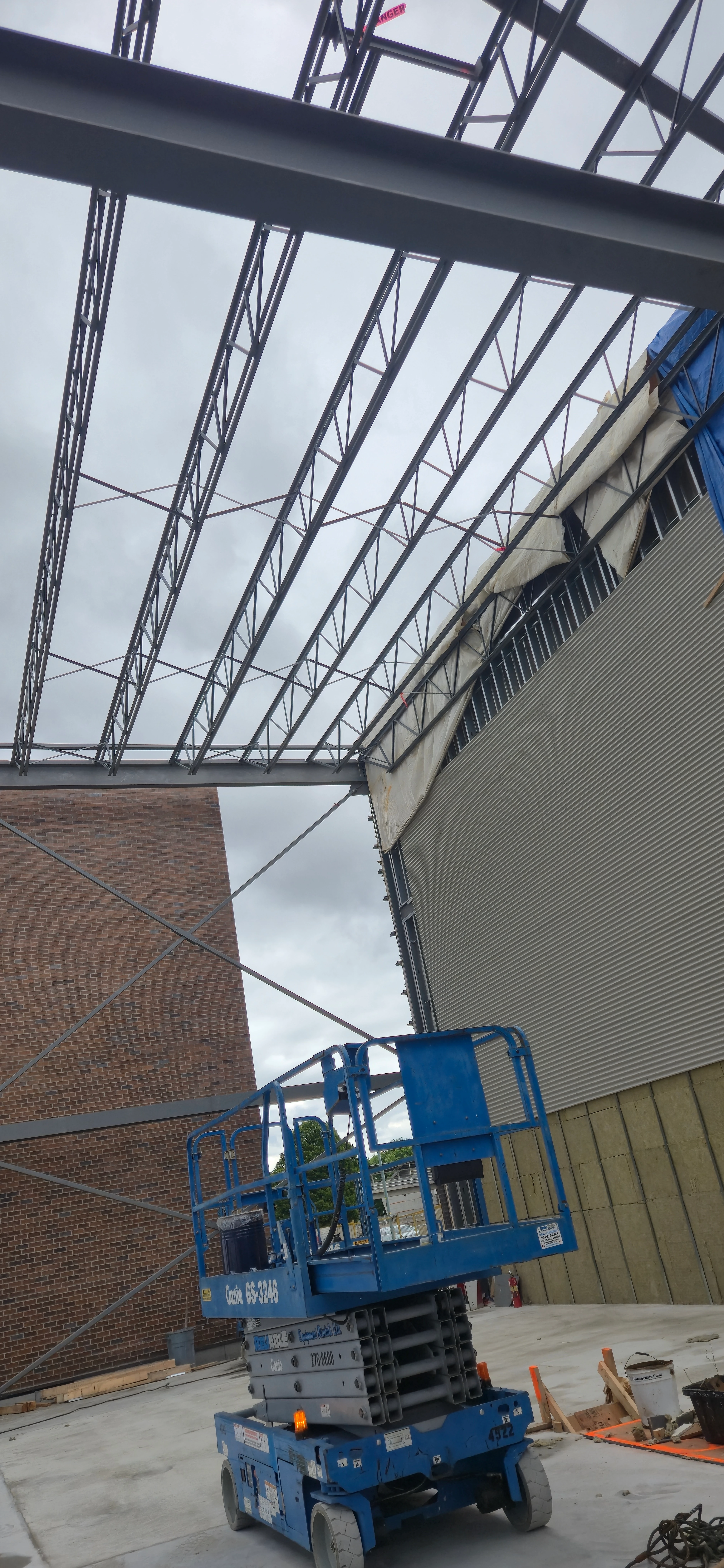 An interior view of a building under construction, looking up at the exposed steel roof joists. A blue scissor lift is parked on the concrete floor below, between a brick wall on the left and an insulated wall on the right.