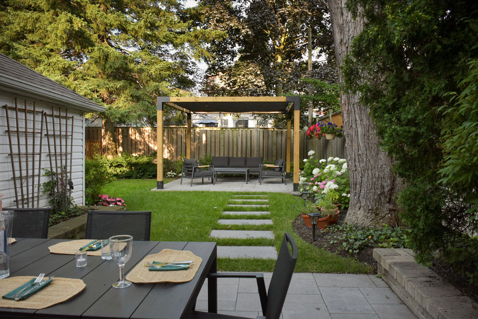 Dinning patio with garden beds on either side looking out towards a stone pathway. The pergola is in the background with planting and tall trees framing the space.