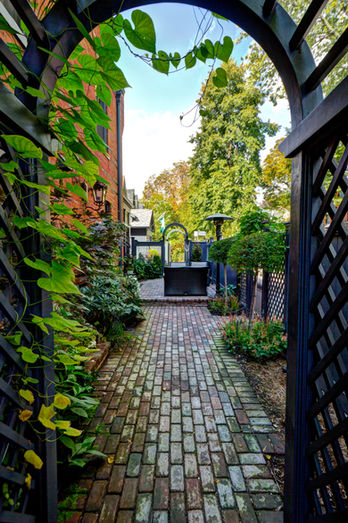 A narrow side yard  with a stone walkway leading to the back. The arbour is dark and has vines growing up it. 