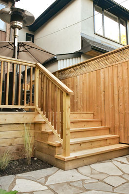 Timber stairs up to a deck from a natural stone patio. To the left of the stairs there is a garden bed with grasses  and to the right a fence sits along the perimeter.