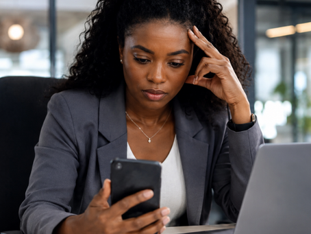 Black woman sitting at her office desk looking concerned while reading work-related messages on her personal cell phone beside an open laptop in a modern workplace setting.