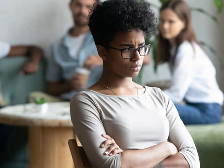 Black woman looking sad and isolated in a workplace setting, with colleagues blurred in the background
