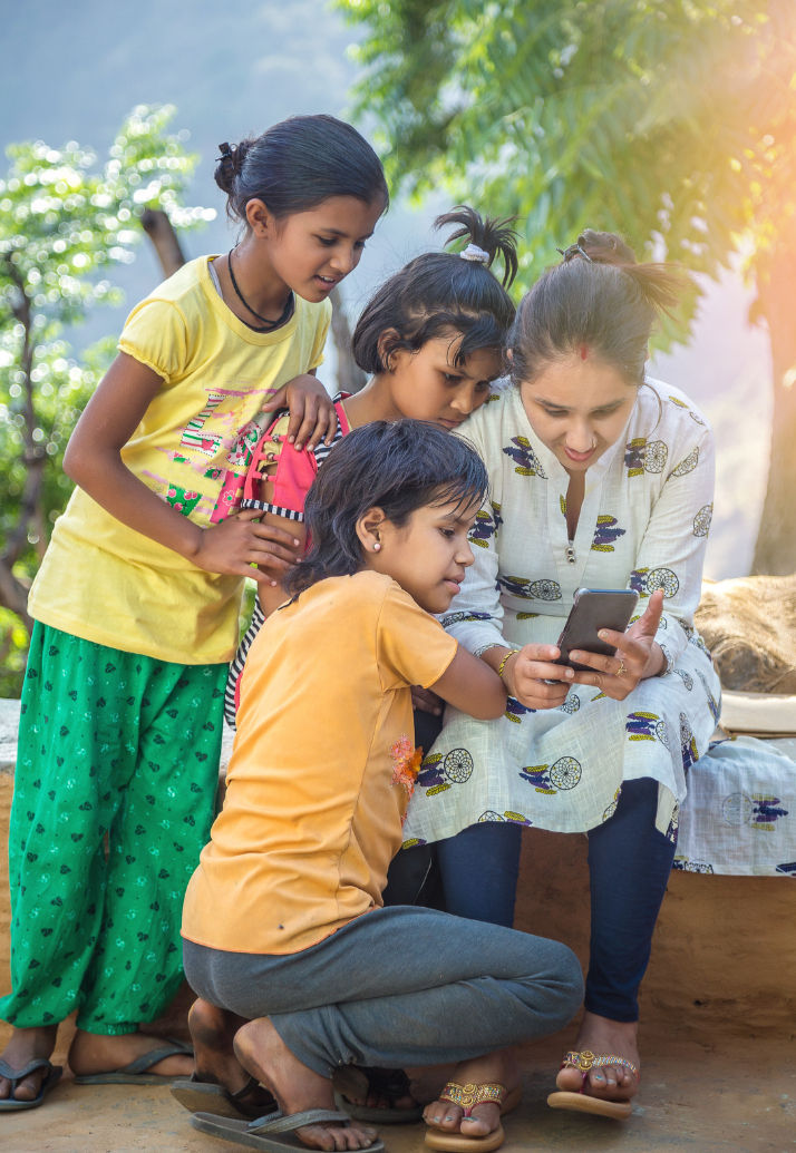 Woman and three children gather around a smartphone, smiling. They're outdoors, surrounded by green trees. Bright, sunny atmosphere.