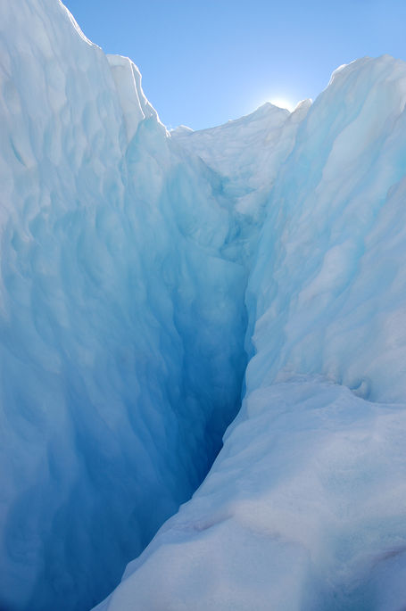 View into a deep blue glacial crevasse enclosed by pale ice walls, with sunlight and blue sky visible through the opening above.