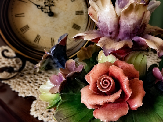 pink and purple porcelain flowers on a doily and a clock in the background with hands at 10:00