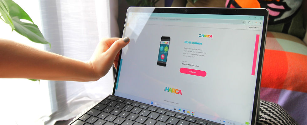 Close-up of a person's hand holding the edge of a laptop displaying the Poplar HARCA "Do it online" digital newsletter ad for online payments and services. The laptop is on someone's lap in a brightly lit room with a plant and a colorful blanket visible in the background. The screen shows information about making payments or accessing services online, optimizing home office technology accessibility and remote service use.