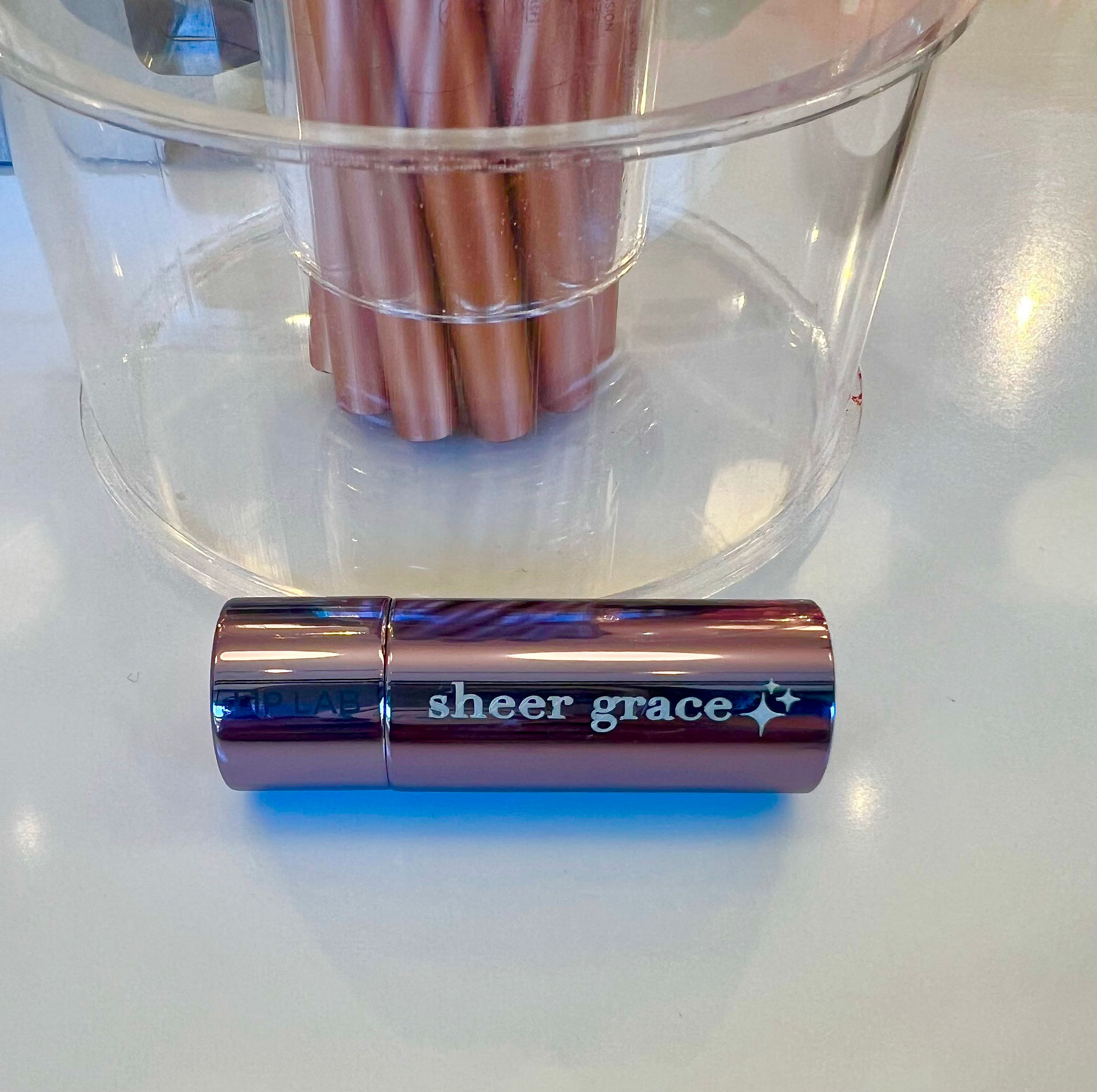 Kimi's custom lipstick tube in a light shade of purple with the words "sheer grace" in all lower case with a sparkle emoji.