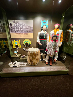 Dance attire and instruments featuring a drum made from hollowed tree trunk and animal hide, cotton fabric clothing on mannequins for a man, a woman, and a child, and head pieces. 
