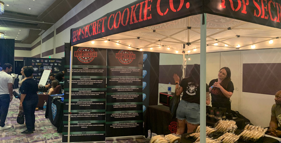 Top Secret Cookie Co. Booth complete with a tent and tables filled with sweet treats and 2 signs displaying the different cookie flavor options.