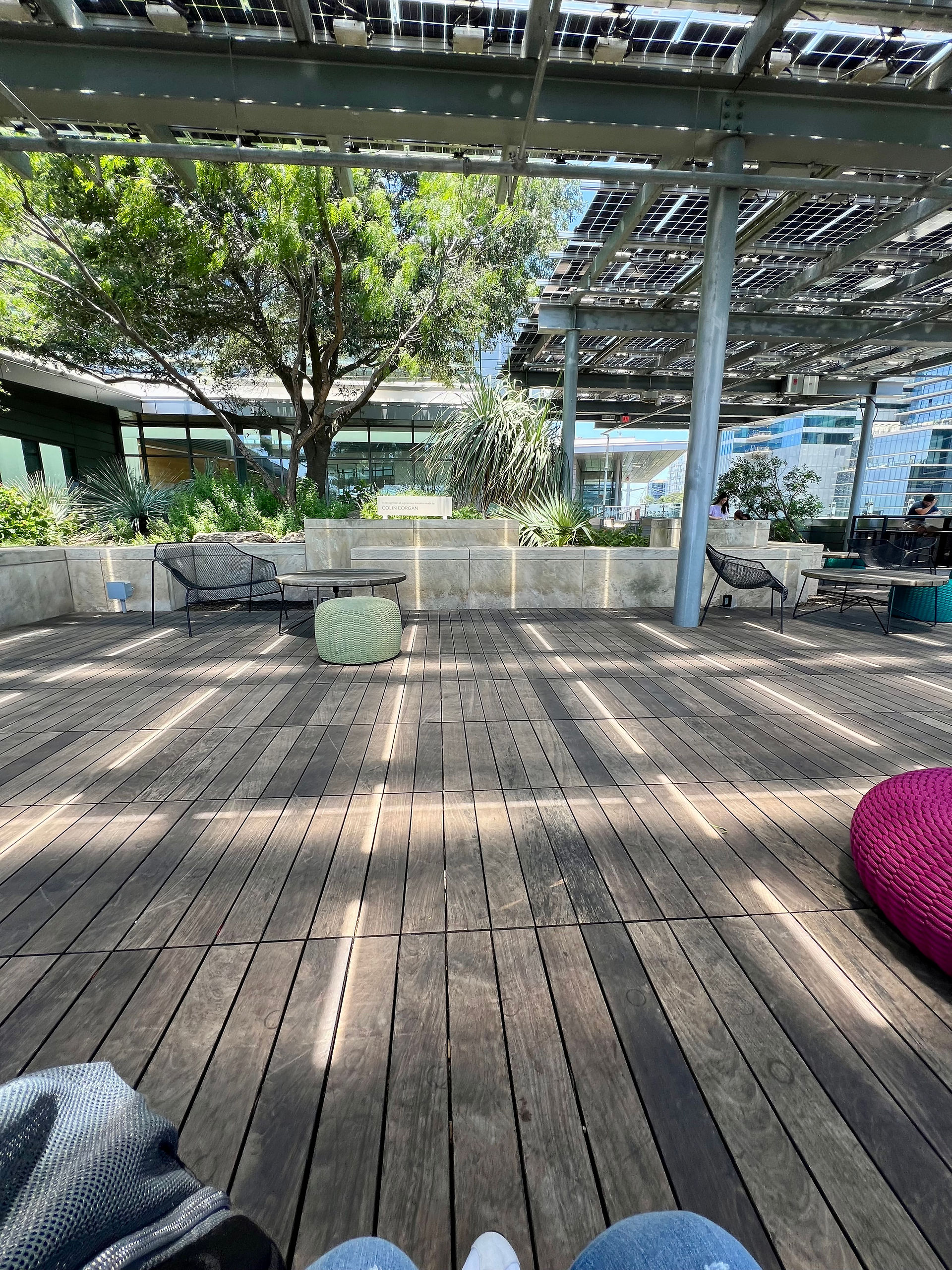 Austin Public Library Rooftop Garden with wooden plank flooring and an awning made from solar panels. There is a butterfly garden with various plants, grasses, and small trees to bring additional life to the outdoor space that has various types of colorful and neutral weatherproof furniture.
