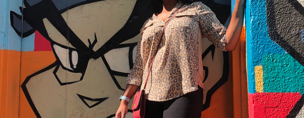 Graffiti Alley picture of Kimi posing in front of a painted bay door in her black jeans and leopard print long-sleeve shirt with black wavy box braids.