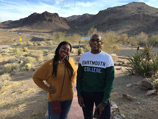 Kimi in a mustard yellow sweater and blue jeans with a small red crossbody purse standing to the left of Mr. Well-Travelled who is wearing a Dartmouth College shirt that is white on the top half and dark green on the bottom. They are standing in front of a desert background at Lake Mead.