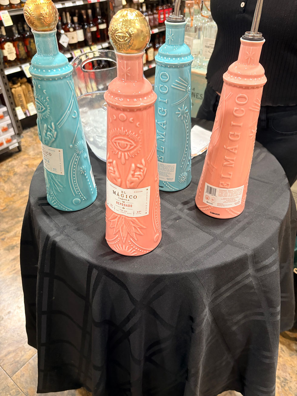 Four pastel blue and pink tequila bottles with gold caps on a black table in a liquor store, labeled El Mágico tequila reposado and El Mágico tequila blanco.