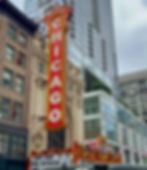 Red vertical Chicago Theater sign with ornate details on a city street, surrounded by tall buildings. Marquee displays show dates.