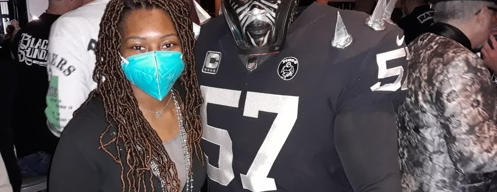Kimi with super fan Violator. Violator is wearing a black Raiders number 57 jersey with silver spikes along with clack and silver chevron face paint and a face shield. Kimi is to his left with a blue N95 mask, gray shirt, black jacket, and Raiders logo beaded necklace. 