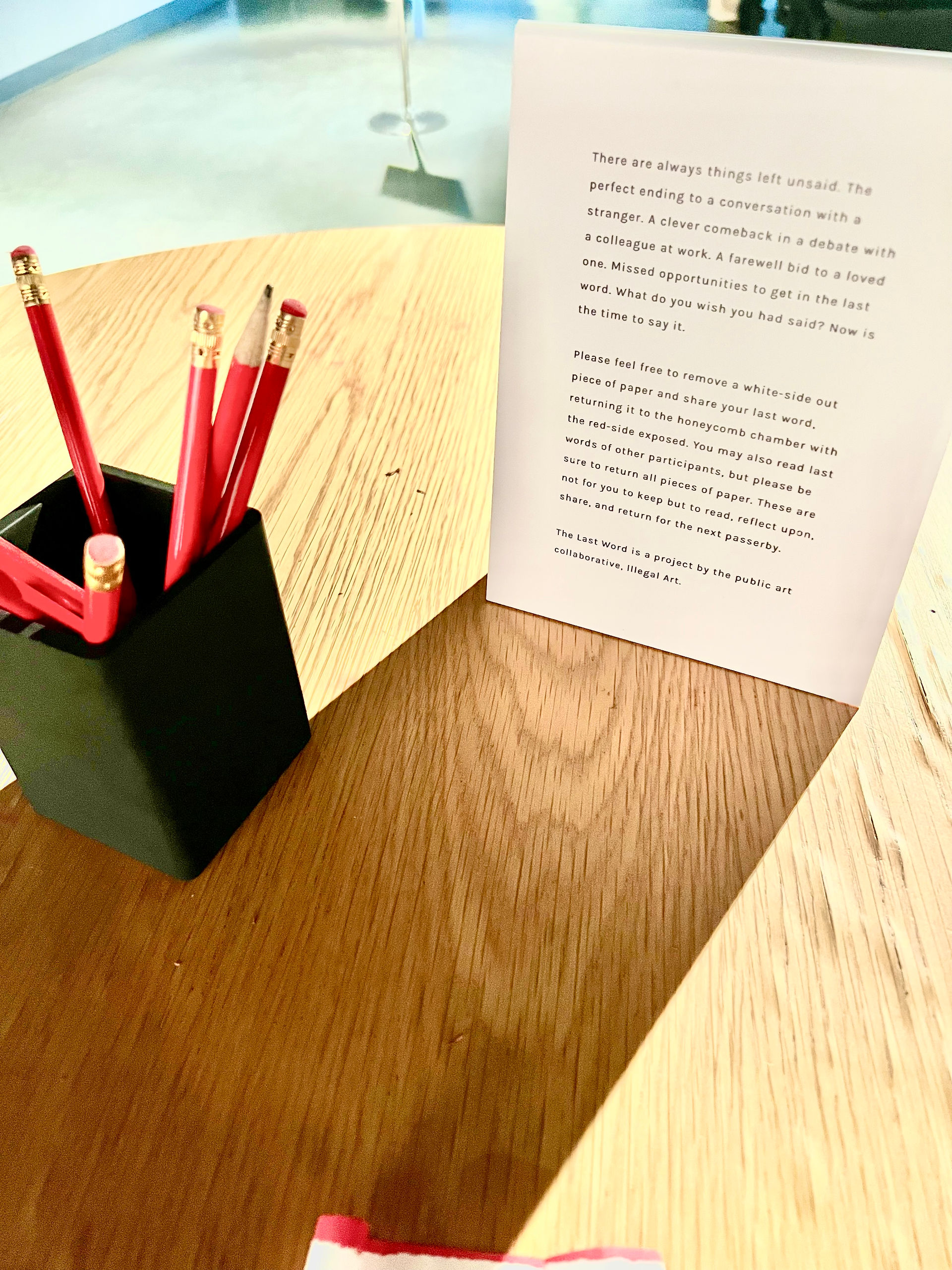 The Last Word by Illegal Art. Image shows a wooden table with a black square container of red wooden pencils and a tabletop placard explaining the purpose of The Last Word and how to participate.