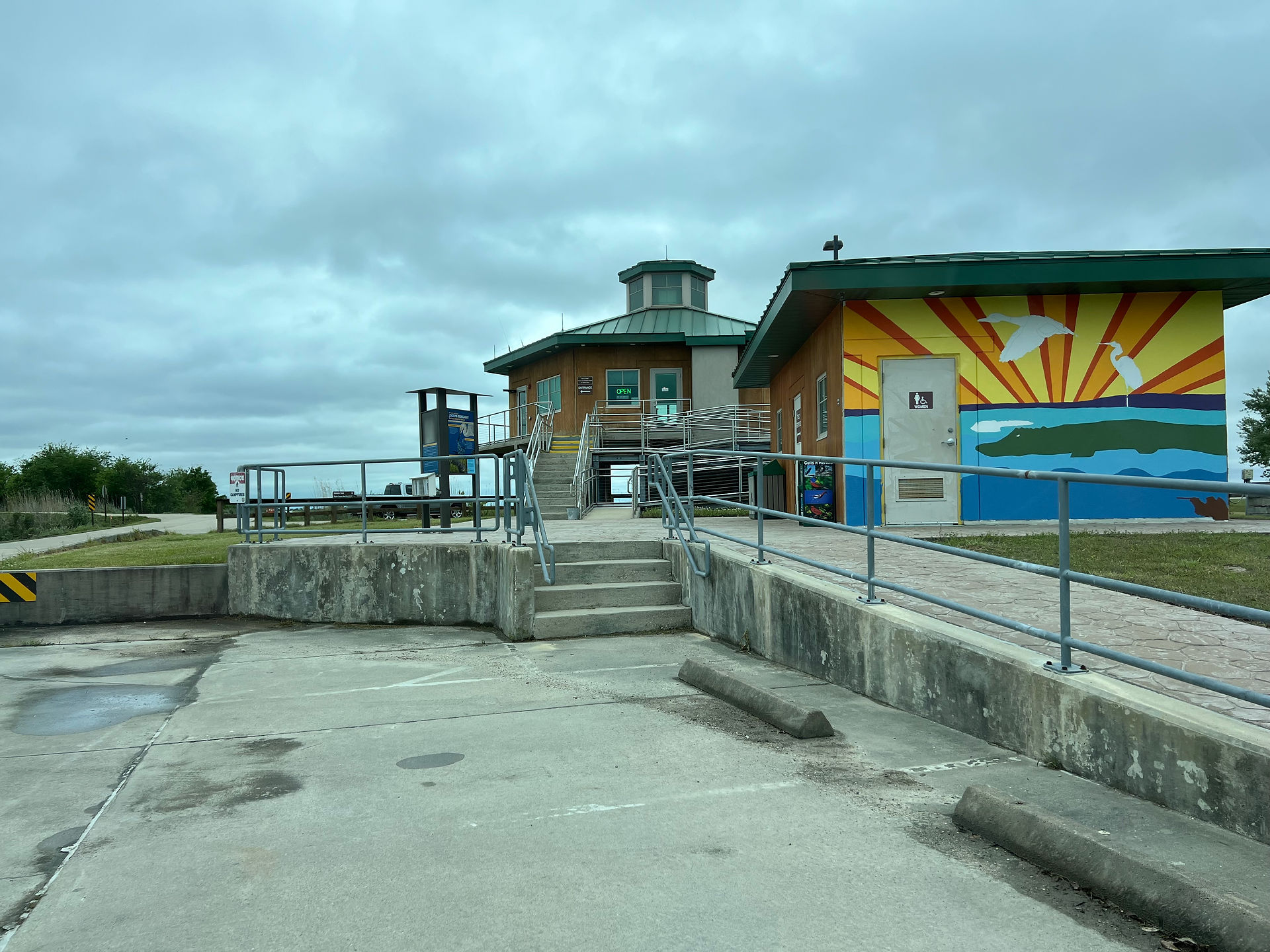 Welcome Center in the shape of a hexagon with a short tower on top. A painted sunrise over the water with a white bird flying across the sky is a mural on the public restrooms to the right of the welcome center. Overcast sky in the background.