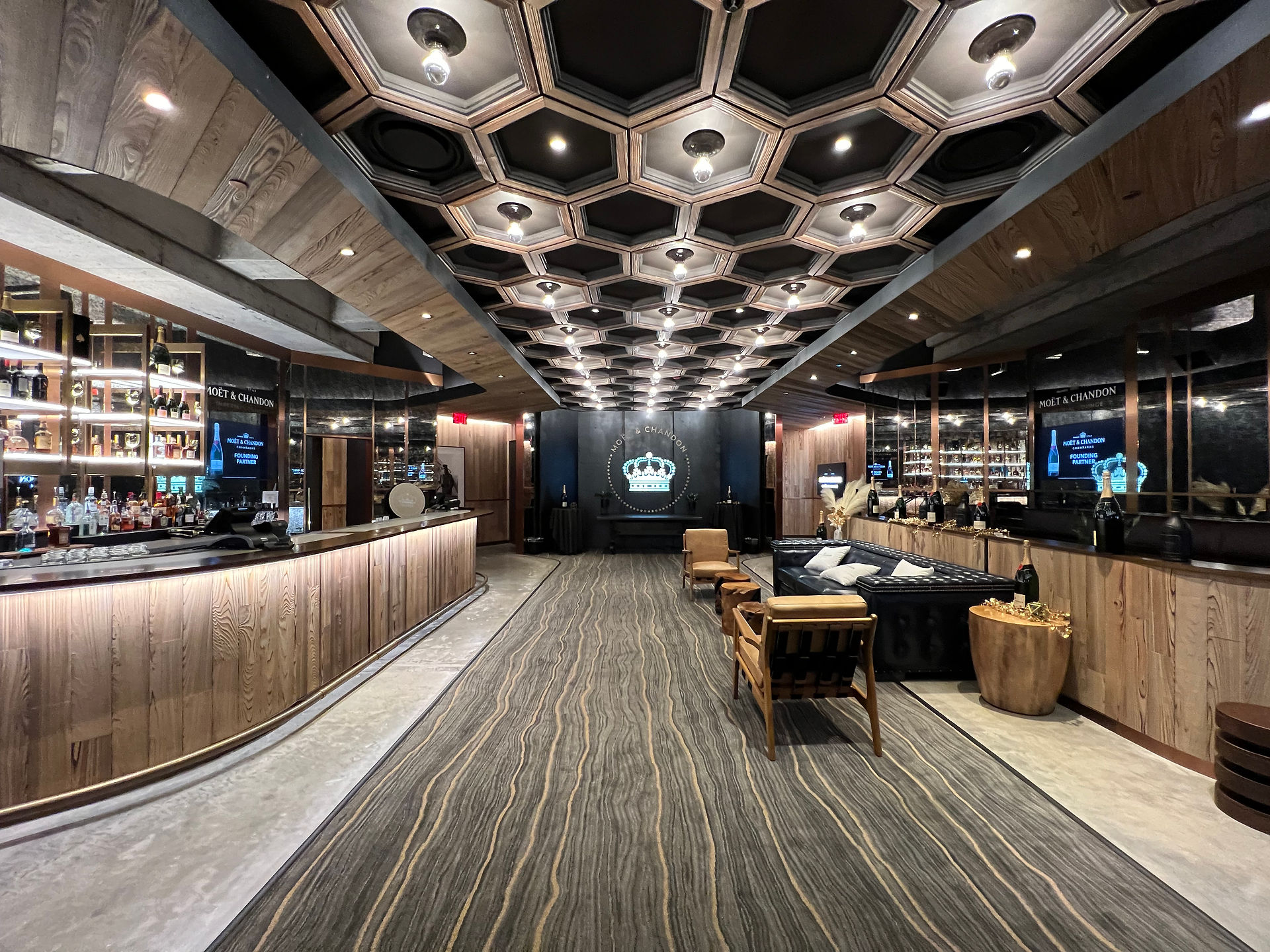 Moët & Chandon Lounge featuring a bar, carpet, wood paneling, comfortable couches and chairs in dark or neutral tones, gold accents, and a honeycomb ceiling
