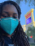 Kimi, a Black woman with brown locks and a blue N95 mask, stands near the Caesars Palace sign in Las Vegas to take a selfie in her black Raiders shirt and silver necklace with her initial. Kimi is to the left of the sign.