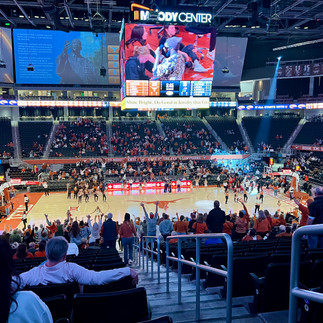 Views of the Court from Section 108 at the Texas Women's Basketball game. Beige colored wood court with burnt orange baselines and a burnt orange longhorn emblem at center court. Above the court is the main scoreboard with the Moody Center name and logo on top. Along the upper walls of the arena are screens with images and information about Martin Luther King, Jr. and the basketball team.