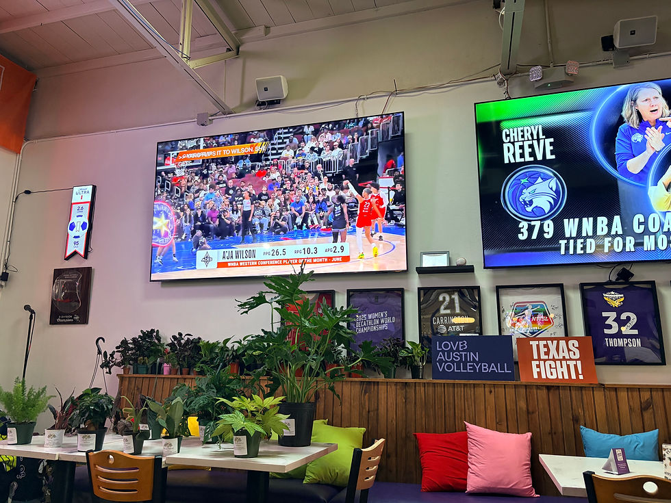 Sports bar with two big TVs showing WNBA highlights, surrounded by potted plants, framed jerseys, and colorful cushions
