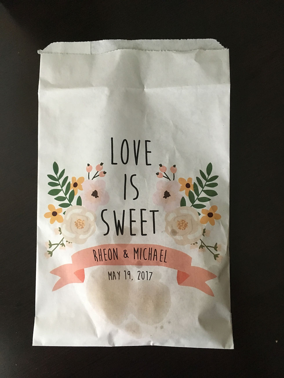 White paper pastry bag with 'Love is Sweet' in black ink, below that is a pink printed ribbon that has the couple's names and their wedding date. The text is framed by pink and yellow flowers with green sprigs to look like the decor.