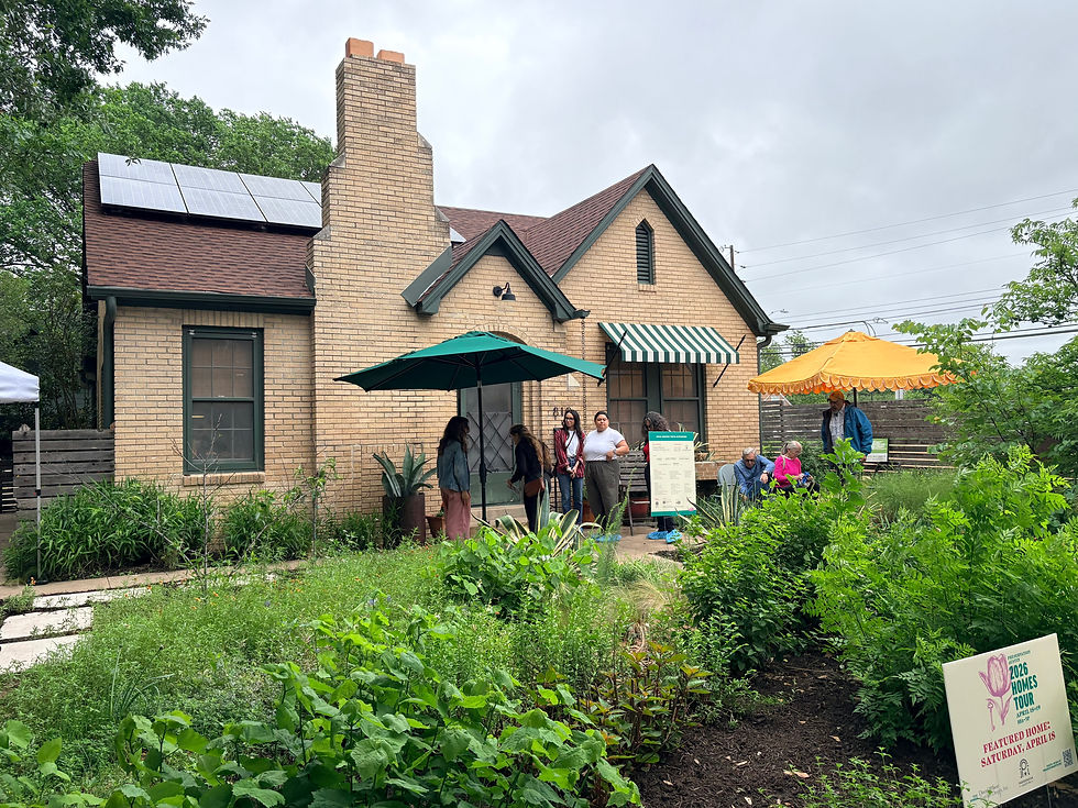 People gather outside a brick house with solar panels, under green and yellow umbrellas. A sign reads "2026 Homes Tour" in a garden setting.
