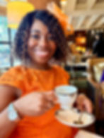 Kimi smiling in an orange dress and matching fascinator holds a teacup in a cozy cafe. Warm lighting, elegant interior, relaxed ambiance.