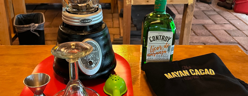 Margarita Prep Bench featuring a blender, margarita glass, handheld citrus juicer, and a jigger are on the left. On the right is a bottle of orange liqueur, and a black apron with The Mayan Cacao Company logo on the front with a white chef's hat underneath the folded apron.