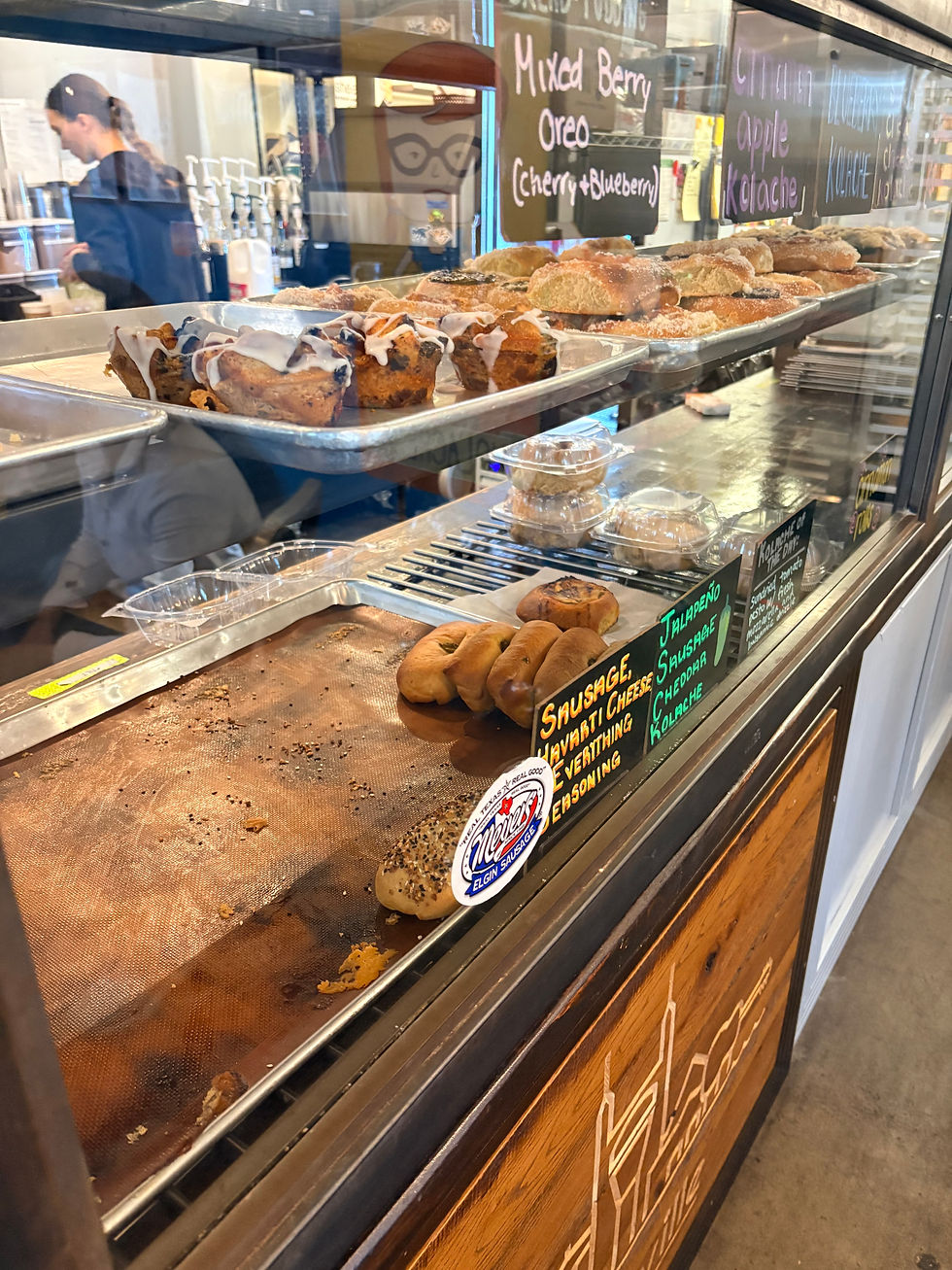 Bakery display with assorted pastries, kolaches, and muffins on trays. Chalkboard highlights flavors. Person works in background. Cozy ambiance.