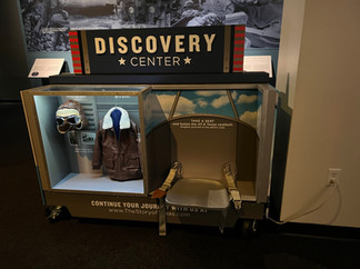 Discovery Center featuring a bomber jacket, hat, and pilot cockpit seat. Children can touch these items and manipulate them.