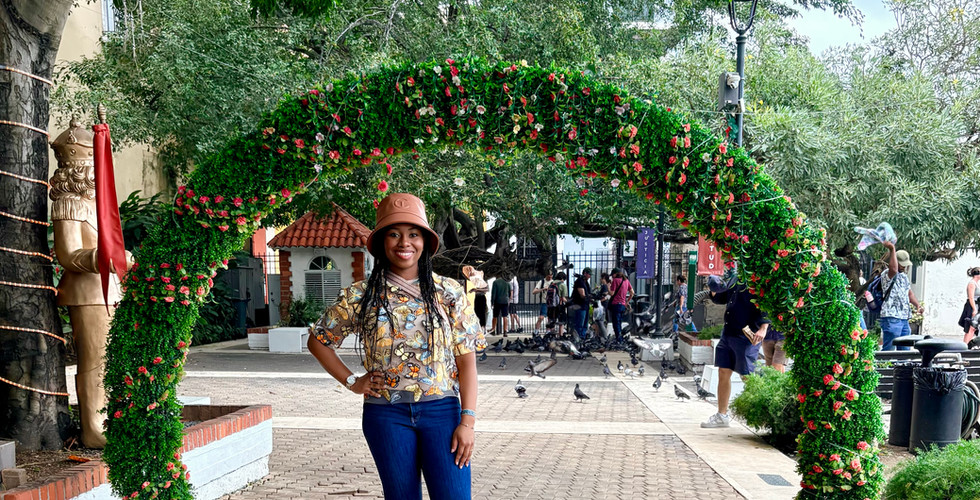 Kimi poses in blue jeans and a sheer top with yellow and blue butterflies on it with a brown bucket hat under a leafy green arch in Parque de las Palomas.