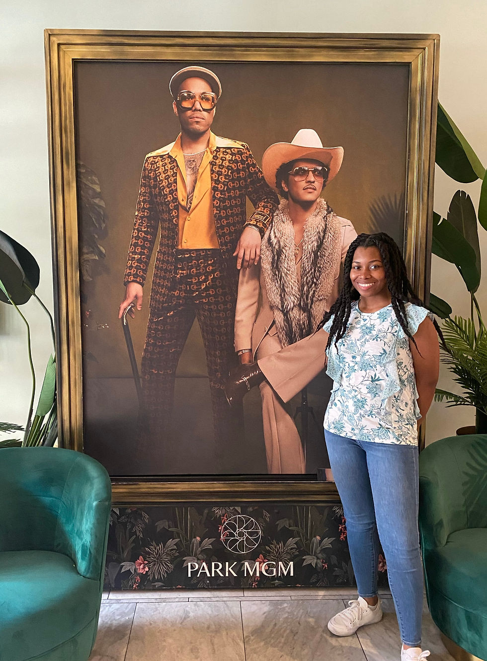 Kimi standing center right of the Silk Sonic poster which is bookended by 2 round, green velvet chairs with large leaf tropical looking plants in the back.
