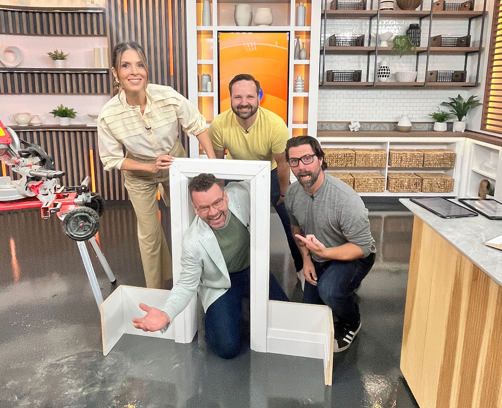Four people smile in a workshop setting with wooden shelves. One crouches in a small frame, pointing. A saw is on the left, creating a playful mood.