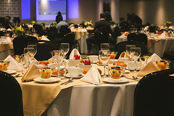 Elegant banquet tables set with fruit cups and glasses, surrounded by seated guests. Blue-lit screen in the background. Warm, formal atmosphere.