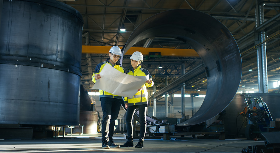 Male and Female Industrial Engineers Look at Project Blueprints While Standing Surround By
