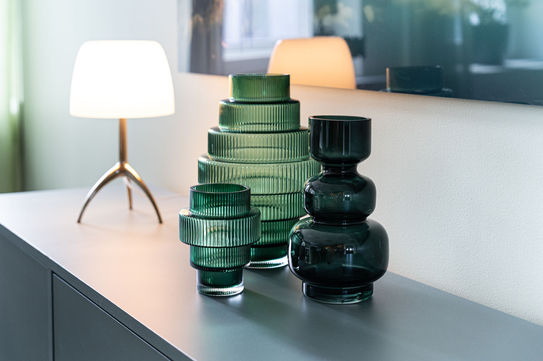 Närbild på grön strukturerad glasvas och en vit designlampa på en ljus möbel i en modern kontorsmiljö.