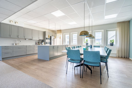 Stort modernt grått kontorspentry med en central köksö och en matgrupp med blå stolar i en öppen planlösning på ett kontor.