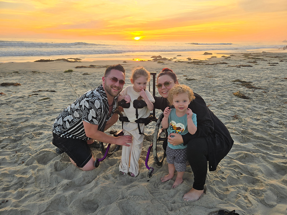 Smiling family of four on a sandy beach at sunset, with one child in a disability assistive device and ocean waves behind them.
