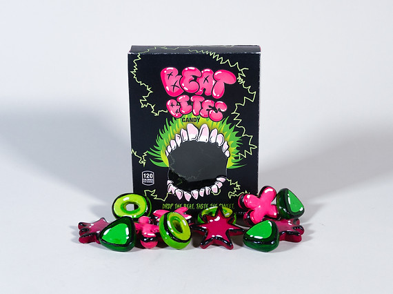 A rectangular black candy box with a mouth opening in the center of the package. The chandy name is located in pink on the front of the box, "beat bites". There is a pile of pink and green candy piled in front of the box on a white surface and backdrop.